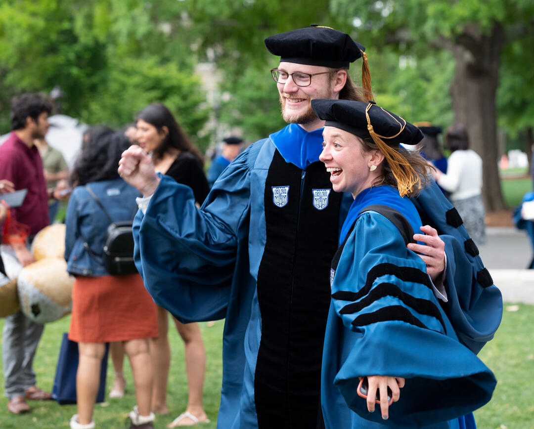 Two graduates celebrating after the 2022 Ph.D. Hooding Ceremony. Both are smiling as they move in to pose for a photo.